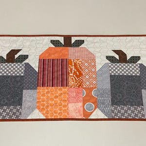 May include: A quilted table runner featuring five stylized pumpkins in shades of orange, gray, and white. The pumpkins have brown stems and are set against a white background with star accents.