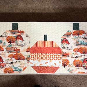May include: A rectangular quilted table runner featuring five pumpkin shapes arranged in a row. Three pumpkins are made of orange fabric strips, while two feature a print of red barns, autumn trees, and vintage trucks on a cream background.
