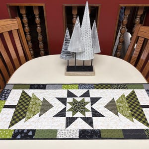 May include: A rectangular quilted table runner featuring a central star motif flanked by two stylized tree shapes. The design incorporates a palette of olive green, black, white, and grey with various geometric and floral patterns in the fabric squares.