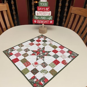 May include: A square Christmas table topper with a star design in the center. The topper is made of red, green, and white patterned fabric squares. A wooden Christmas tree decoration with the text "May Your Days Be Merry and Bright" sits on the table.