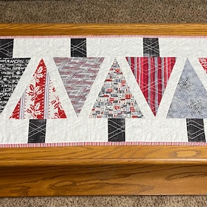 May include: A rectangular Christmas table runner featuring six stylized Christmas trees in red, black, and gray patterns. The trees are set against a white background with a red and white striped border. Two star shapes are on the ends.