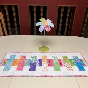 May include: A rectangular quilted table runner featuring a white background with wavy lines and a series of vertical fabric rectangles in various colors and patterns, accompanied by a metal flower sculpture with a green stem and base.