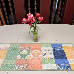 May include: A rectangular quilted table runner featuring a patchwork design of various fabrics in shades of green, blue, orange, and yellow, with patterns including floral prints and chickens, finished with a thin green border trim.