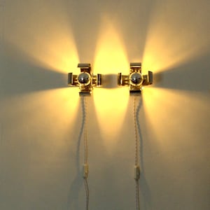 May include: Two metallic wall-mounted lamps with a cross-shaped design, each emitting a bright, warm light. The fixtures feature a central bulb and are connected to white power cords with inline switches hanging vertically against the wall.