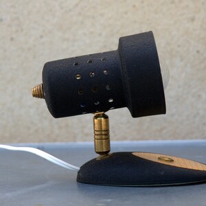 May include: A black and brass desk lamp with a round, perforated shade and a white cord. The lamp has a small, round base with a wooden accent.