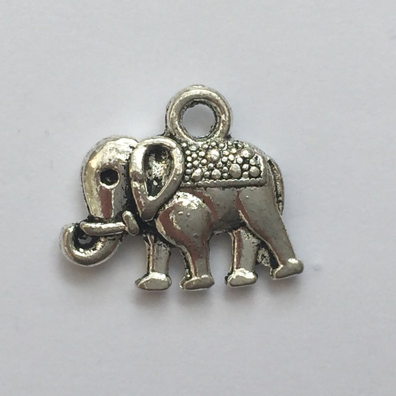 May include: A small, silver-colored elephant charm with a loop at the top for attaching to jewelry or other crafts. The elephant has detailed features, including a textured saddle and trunk. The charm is made of metal and has a vintage or antique appearance.