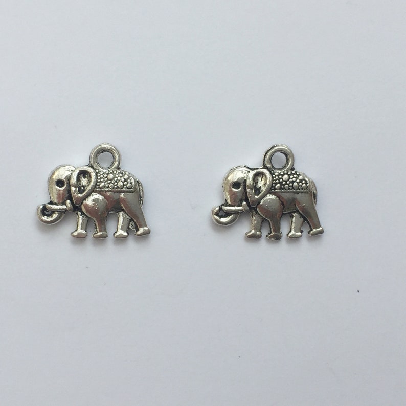 May include: Two small, silver-colored elephant charms. Each charm features a detailed elephant design with a textured back and a loop at the top for attaching to jewelry or other crafts. The charms are displayed against a white background.