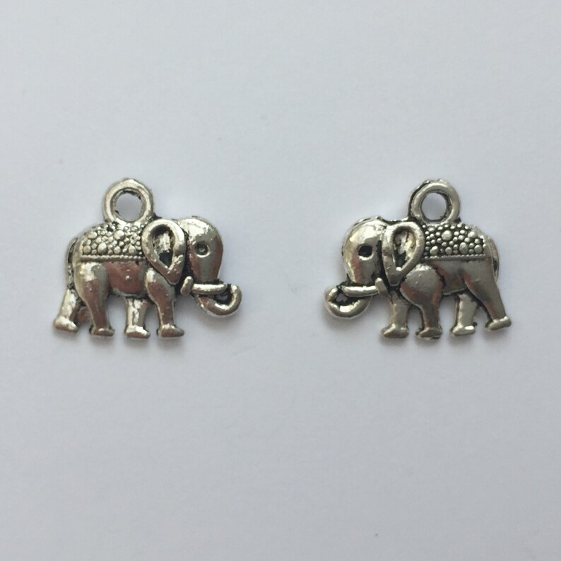 May include: Two silver-colored elephant charms. Each charm features an elephant with detailed trunk, ears, and a textured blanket design on its back. The charms have a small loop at the top for attaching to jewelry or other crafts.