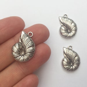 May include: Three silver-toned metal charms shaped like seashells. The shells have a detailed, textured design and a loop at the top for attaching to jewellery.