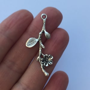 May include: Silver charm featuring a stylised branch with leaves and a flower. The charm has a loop at the top for attaching to a necklace or bracelet.