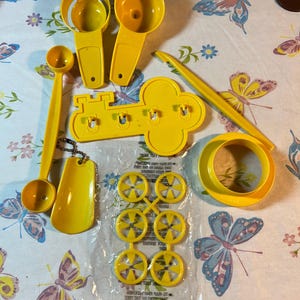 May include: A set of yellow plastic kitchen tools, including a funnel, a scoop, a key-shaped tool, a ring mold, and a set of six star-shaped cutters. The tools are packaged in a clear plastic bag with instructions.