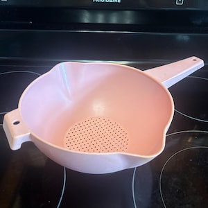 May include: A light pink plastic colander with a long handle and a small side tab, both featuring circular holes for hanging. The bowl has a perforated base for draining and a molded pouring spout on the side, designed for kitchen food preparation.