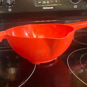 May include: A bright red plastic colander featuring a wide, rounded bowl shape, a long handle on one side, and a small tab handle on the opposite side. The base of the colander includes a perforated section for draining liquids during food preparation.