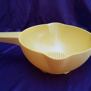 May include: A light yellow plastic colander with a long handle and a pouring spout. The colander has a bowl shape with small holes at the bottom for draining liquids. It is designed for kitchen use.