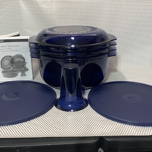 May include: A dark blue Tupperware Stack Cooker set with multiple components, including a stackable pot, lids, and a central stand. The set includes an instruction manual. The items are arranged on a white surface.