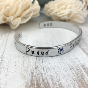 May include: Silver cuff bracelet with the words "Proud Police" and a blue police badge charm.