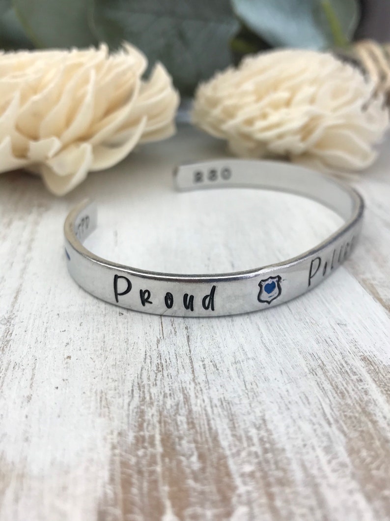 May include: Silver cuff bracelet with the words "Proud Police" and a blue police badge charm.