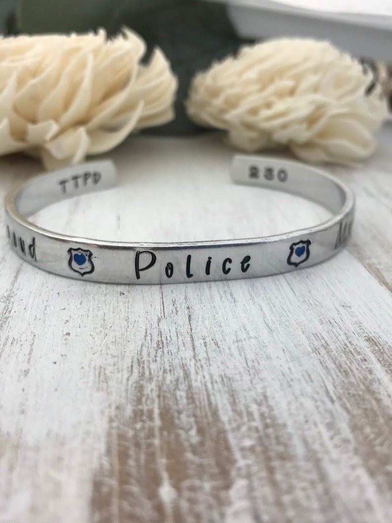 May include: A silver bracelet with the words "Police" and "TTPD" engraved on it. The bracelet also has two small blue badges with a heart in the center.