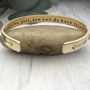 May include: Gold bangle bracelet with an inscription that reads "Beautiful girl, you can do hard things".