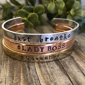 May include: Three stacked metal bracelets with engraved text. The top bracelet is silver and says "just breathe". The middle bracelet is rose gold and says "#LADY BOSS". The bottom bracelet is gold and says "#Queenbee".