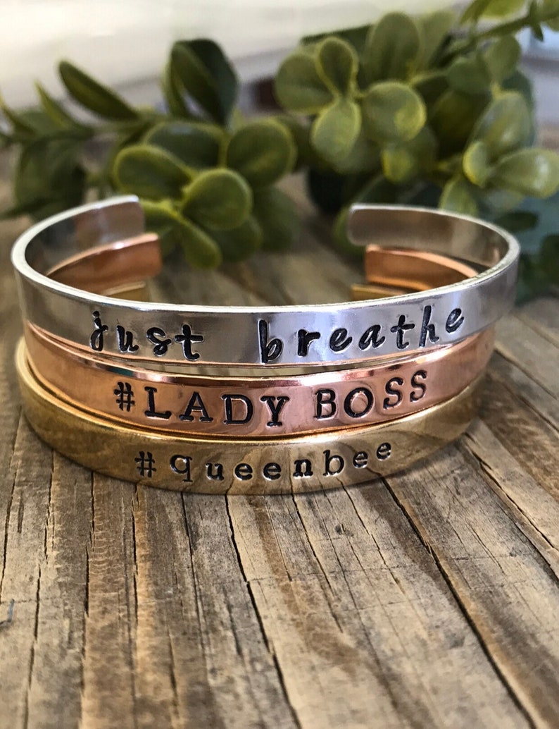 May include: Three stacked metal bracelets with engraved text. The top bracelet is silver and says "just breathe". The middle bracelet is rose gold and says "#LADY BOSS". The bottom bracelet is gold and says "#Queenbee".