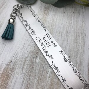 May include: A silver bookmark with a floral design and the text "Just one more chapter" with a heart. The bookmark has a teal tassel.