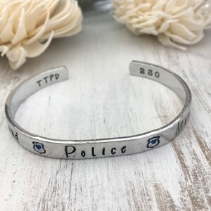 May include: Silver bracelet with the words "TTPD 230" and "Police" engraved on it. The bracelet also has a small blue enamel badge with a heart.