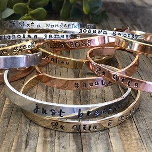 May include: A stack of silver and gold bracelets with various engraved sayings, including "Lady Boss", "Just Breathe", "La Vita e Bella", and "#hust".