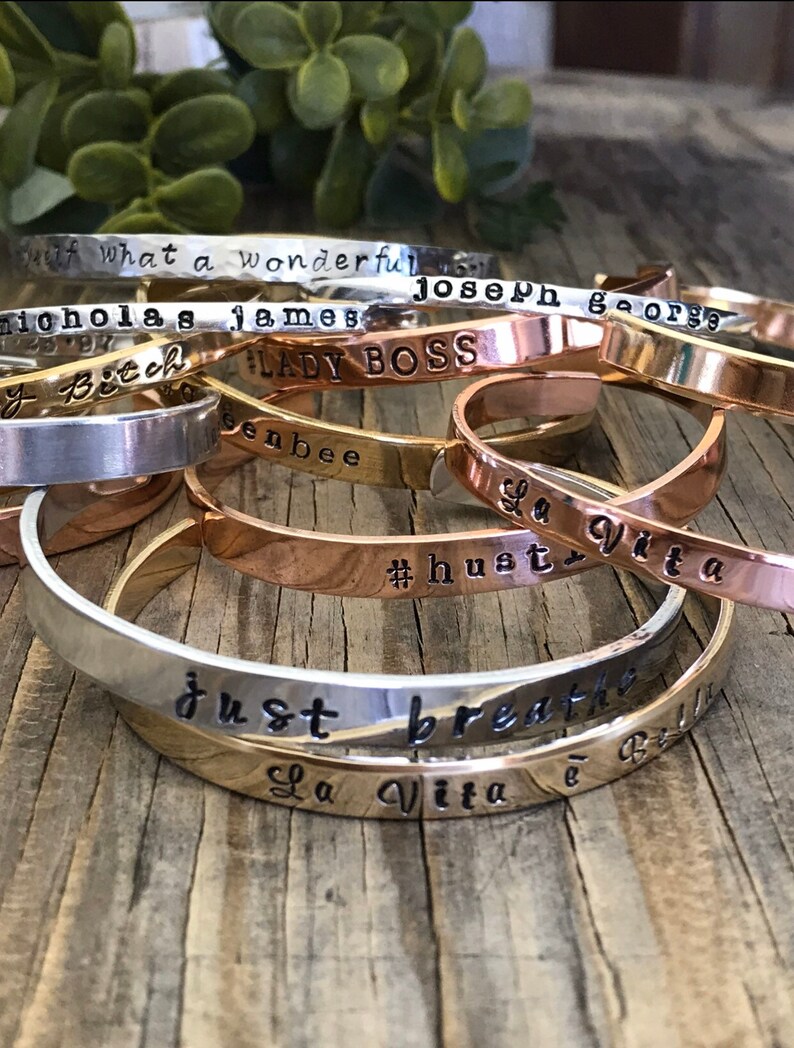 May include: A stack of silver and gold bracelets with various engraved sayings, including "Lady Boss", "Just Breathe", "La Vita e Bella", and "#hust".