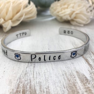 May include: Silver bracelet with the words "Police" and "My" engraved on it. The bracelet also has a small blue badge charm with a heart in the center.