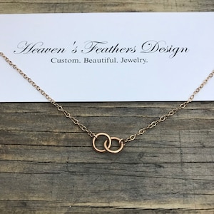 May include: A gold chain necklace with two interlocking circles as a pendant. The necklace is on a wooden surface with a white card that reads "Heaven's Feathers Design Custom. Beautiful. Jewelry."