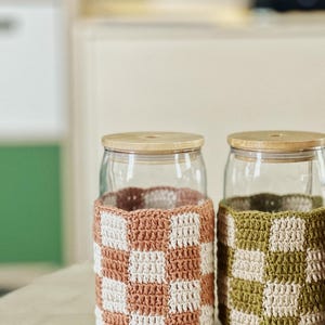 May include: Two glass jars with wooden lids, each wrapped with a crocheted checkerboard pattern. One jar has a pink and white design, the other has a green and white design. The jars sit on a light-coloured surface.