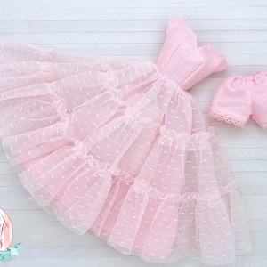 May include: A light pink doll dress and matching shorts set. The dress has a tiered skirt with a dotted overlay and a fitted bodice. The shorts have a lace trim. The set is designed for dolls.