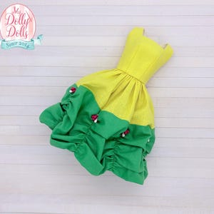 May include: A doll dress in bright yellow and green. The bodice is yellow, and the skirt is divided into yellow and green sections, with gathered ruffles. Small red and white accents are visible. The dress is on a white wooden surface. The logo says "My Dolly Dolls Since 2014".