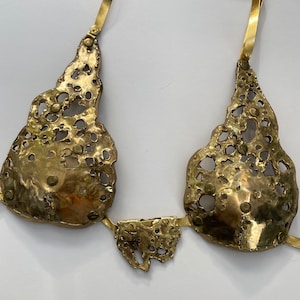 Op de afbeelding: Een gouden metalen ketting met een uniek, abstract ontwerp. De ketting heeft twee grote, opengewerkte stukken die met elkaar verbonden zijn door een kleiner, rechthoekig stuk. De ketting is gemaakt van een getextureerd metaal met een gehamerde afwerking.
