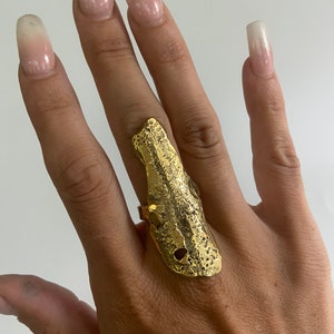 Puede incluir: Un anillo abierto de textura dorada con una forma única e irregular. El anillo se lleva en el dedo índice de una mano.