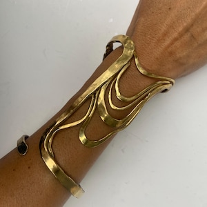 Puede incluir: Un brazalete de color dorado con un diseño abstracto y fluido. El brazalete se envuelve alrededor del brazo, con múltiples capas curvas y un diseño abierto. La joya es de metal y tiene una textura martillada.