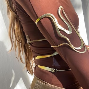 May include: A brown long-sleeve top and gold-colored pants are paired with a gold arm cuff. The arm cuff has a unique, flowing design. The outfit is a stylish ensemble.