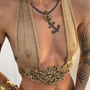Könnte beinhalten: Goldfarbenes Metall-Haltertop mit Kettengeflecht-Design. Das Top hat ein dekoratives, strukturiertes Band an der Taille. Ein goldener Choker und eine Halskette mit Anhänger sind ebenfalls zu sehen. Körperschmuck und Accessoires.