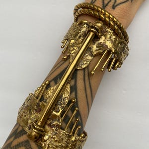 Puede incluir: Un brazalete de metal dorado con un diseño texturizado y orgánico. El brazalete presenta una barra central con una parte superior redondeada y una serie de pequeñas protuberancias redondeadas a lo largo de los bordes.