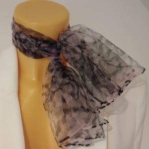 Könnte beinhalten: Ein transparenter Schal mit einem schwarzen und lavendelfarbenen Muster ist um den Hals einer Schaufensterpuppe drapiert. Der Schal hat einen gerüschten Rand und besteht aus einem leichten, fließenden Stoff. Der Hintergrund ist eine weiße Oberfläche.