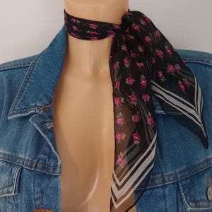 Könnte beinhalten: Ein schwarzer Schal mit einem Muster aus rosa Rosen und grünen Blättern. Der Schal ist um den Hals einer Schaufensterpuppe drapiert, über einer Jeansjacke. Der Schal hat weiße und schwarze Streifen an den Rändern.