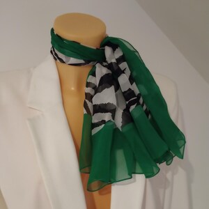 May include: A sheer scarf with a green border and a black and white zebra stripe pattern. The scarf is draped around a mannequin's neck, with the green border cascading down. The scarf is paired with a white blazer.
