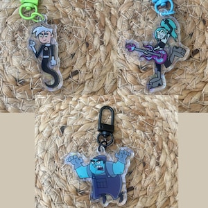 May include: Three clear plastic keychains featuring characters from the cartoon series Danny Phantom. The first keychain features Danny Phantom, a boy with white hair and a black and white ghost costume. The second keychain features a girl with green hair and a black and purple outfit playing a guitar. The third keychain features a blue ghost with a black and blue outfit.