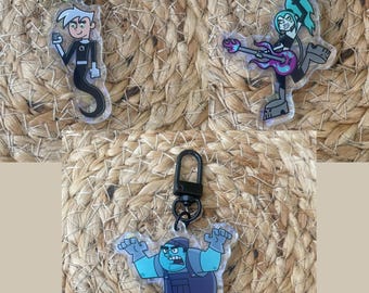 May include: Three clear plastic keychains featuring characters from the cartoon series Danny Phantom. The first keychain features Danny Phantom, a boy with white hair and a black and white ghost costume. The second keychain features a girl with green hair and a black and purple outfit playing a guitar. The third keychain features a blue ghost with a black and blue outfit.