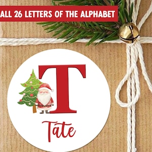 Puede incluir: Una pegatina redonda blanca con la letra "T" roja y el nombre "Tate" en escritura roja. La pegatina presenta una ilustración de un árbol de Navidad y Papá Noel. El texto "ALL 26 LETTERS OF THE ALPHABET" está en la parte superior.