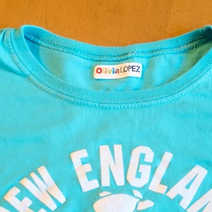 May include: A turquoise blue t-shirt with a white graphic that says "NEW ENGLAND" and a white image of a lobster. The shirt has a white label sewn into the neckline that says "Olivia LOPEZ".