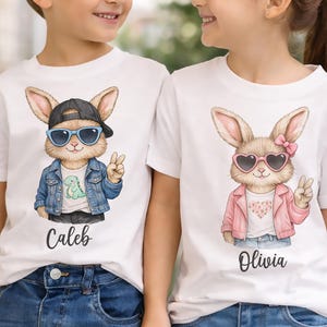 Könnte beinhalten: Weiße T-Shirts mit Cartoon-Hasen. Ein Hase trägt eine schwarze Kappe, blaue Sonnenbrille und eine Jeansjacke. Der andere Hase trägt herzförmige Sonnenbrillen und eine rosa Jacke. Die Namen "Caleb" und "Olivia" sind aufgedruckt.
