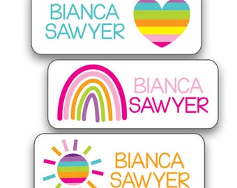 Puede incluir: Tres etiquetas termoadhesivas coloridas con el nombre "Bianca Sawyer" y diferentes diseños. La primera etiqueta tiene un corazón arcoíris, la segunda un arcoíris y la tercera un sol.