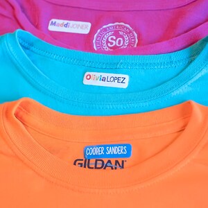 May include: Three colorful iron-on labels with names on them. The top label says "Maddi JOINER", the middle label says "Olivia LOPEZ", and the bottom label says "COOPER SANDERS". The bottom label also has the word "GILDAN" printed on it.